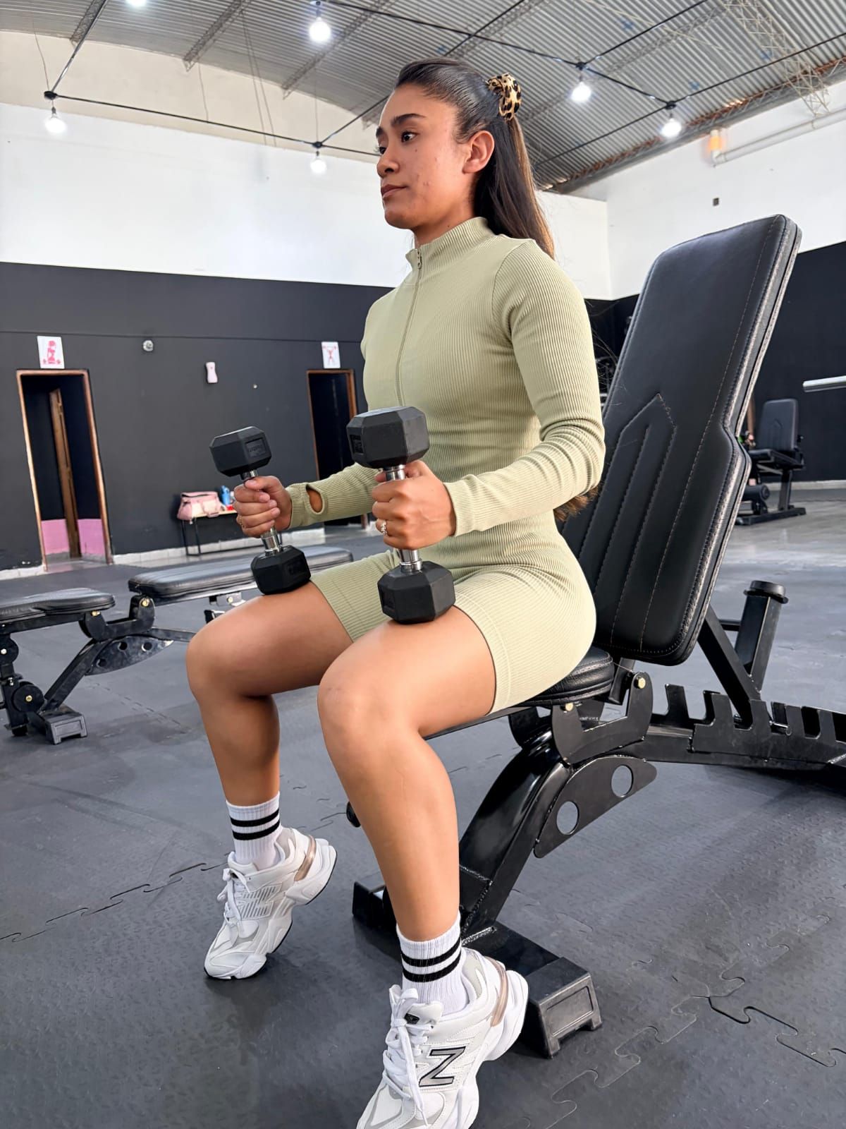 Persona sentada en un banco de gimnasio sosteniendo mancuernas, vestida con ropa deportiva verde y zapatillas blancas.