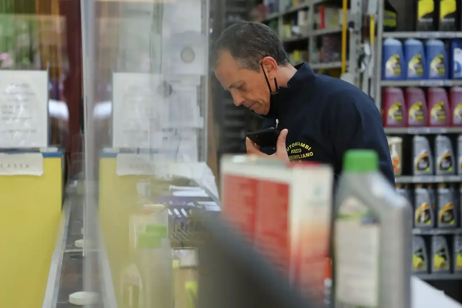 Un uomo in camicia blu osserva un articolo dietro un bancone trasparente, all'interno di un negozio. Sullo sfondo, scaffali di bottiglie.