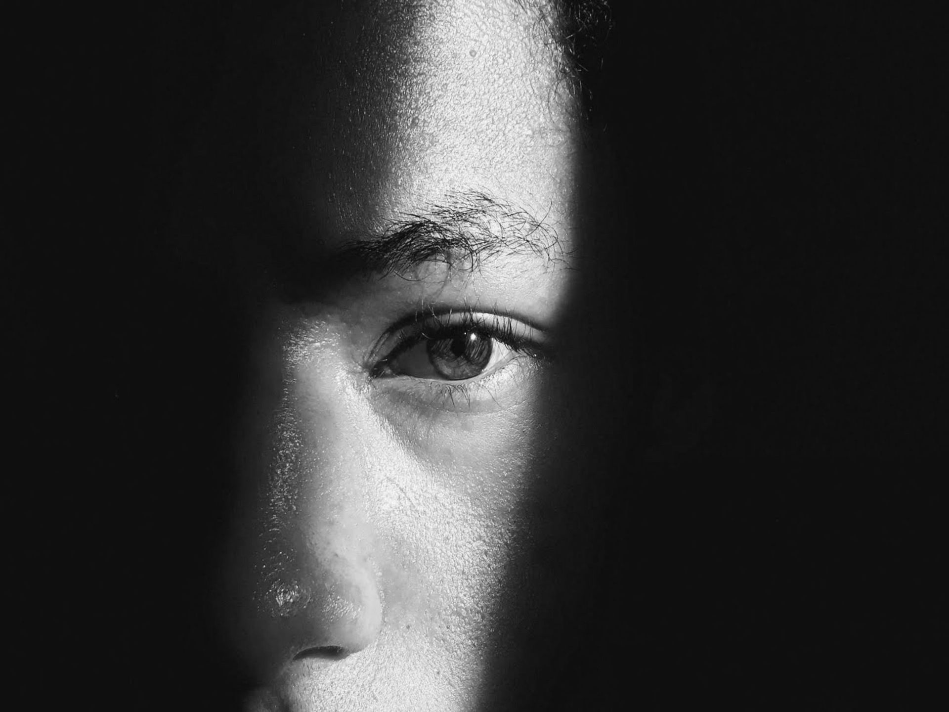 Close-up of a face in shadow, with one eye visible and a bright light across the forehead