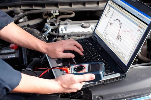 A man is working on a laptop computer under the hood of a car