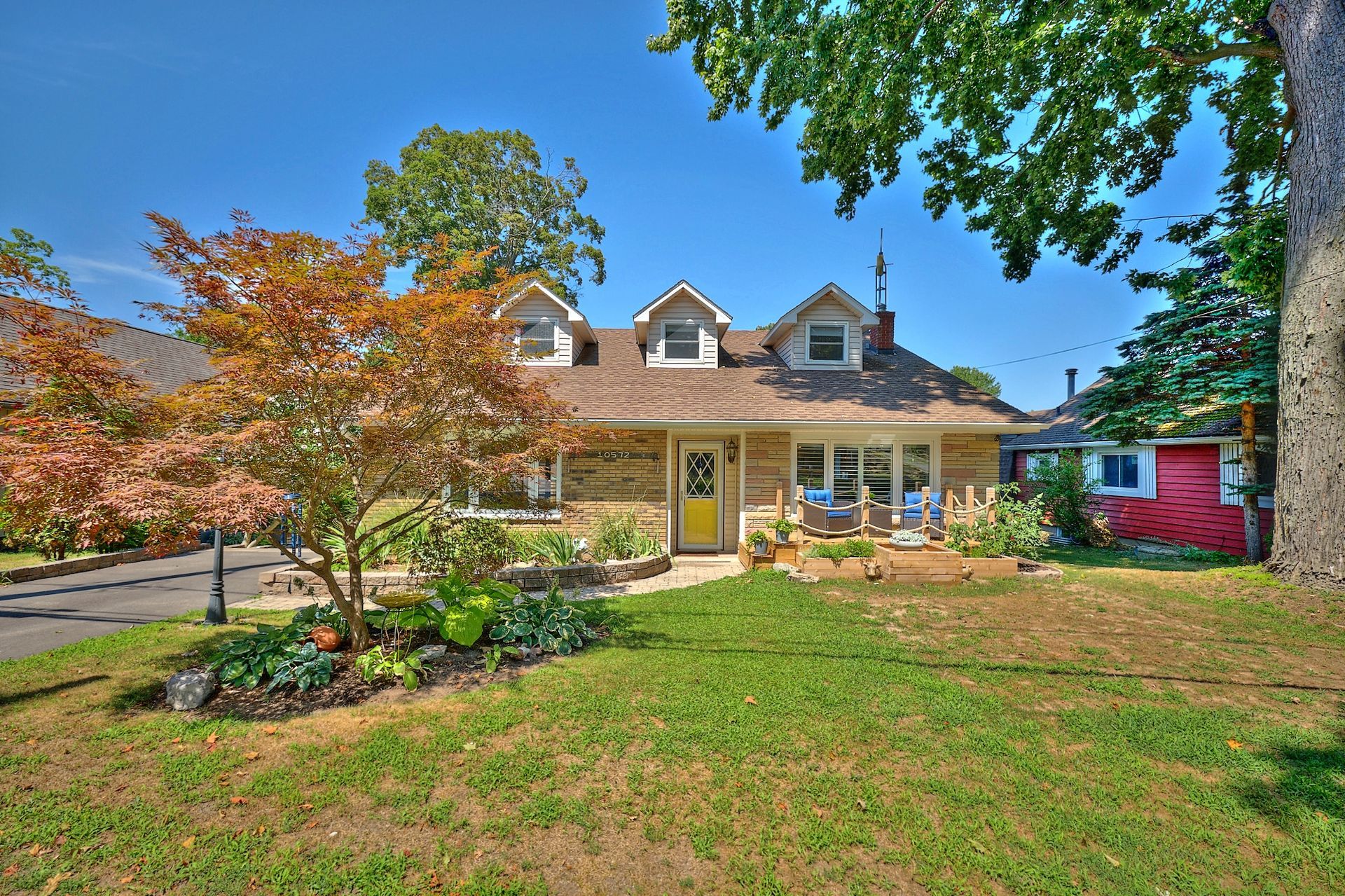 Front exterior of a two-storey home with windows, a yellow front door, and landscaped garden beds under a bright blue sky.
