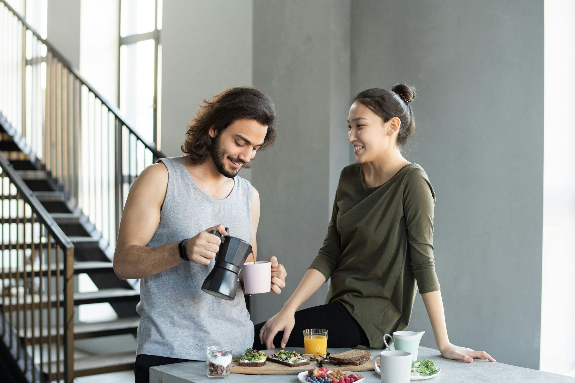 Man pouring coffee for a woman, both smiling near a table of breakfast food. Indoor setting.