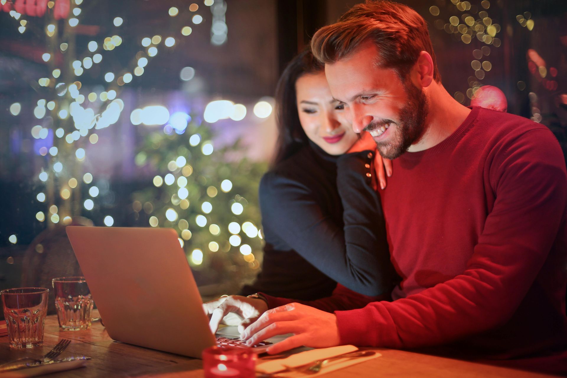 Couple smiling, looking at laptop in a restaurant with string lights. Man types, woman rests hand on his shoulder.