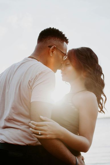Couple embracing, faces close together, with sunlight between them. One wears glasses, other has engagement ring.