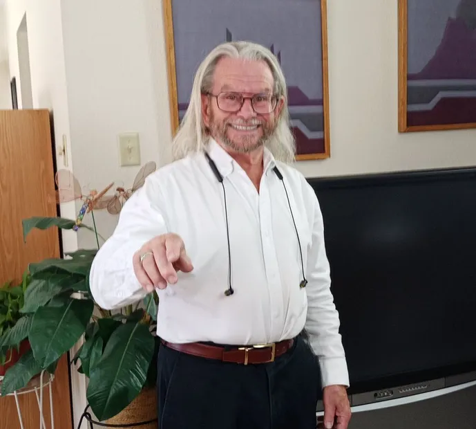 Man with long white hair, glasses, white shirt, points towards the viewer, smiling in a room.