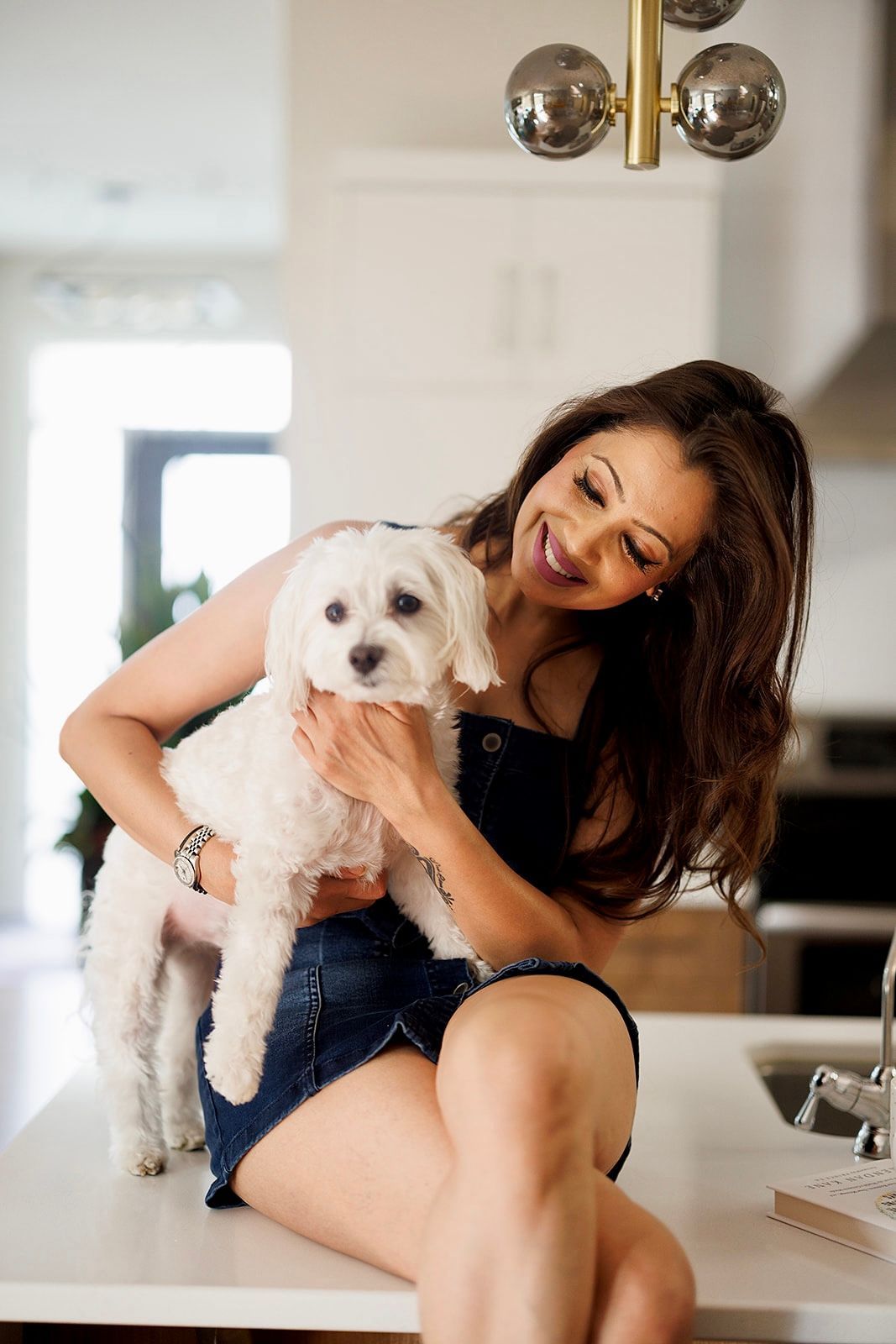 A woman is sitting on a counter holding a small white dog.