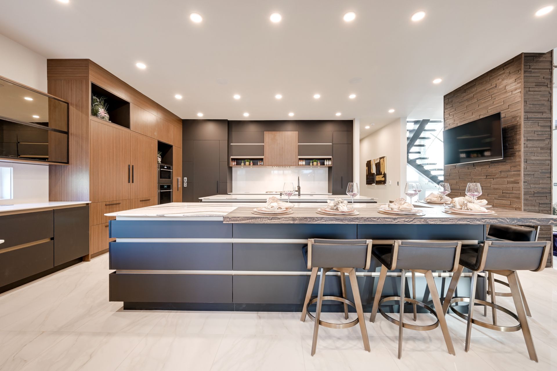 Modern kitchen with large island, dark cabinets, wood accents, and seating; TV on brick wall.