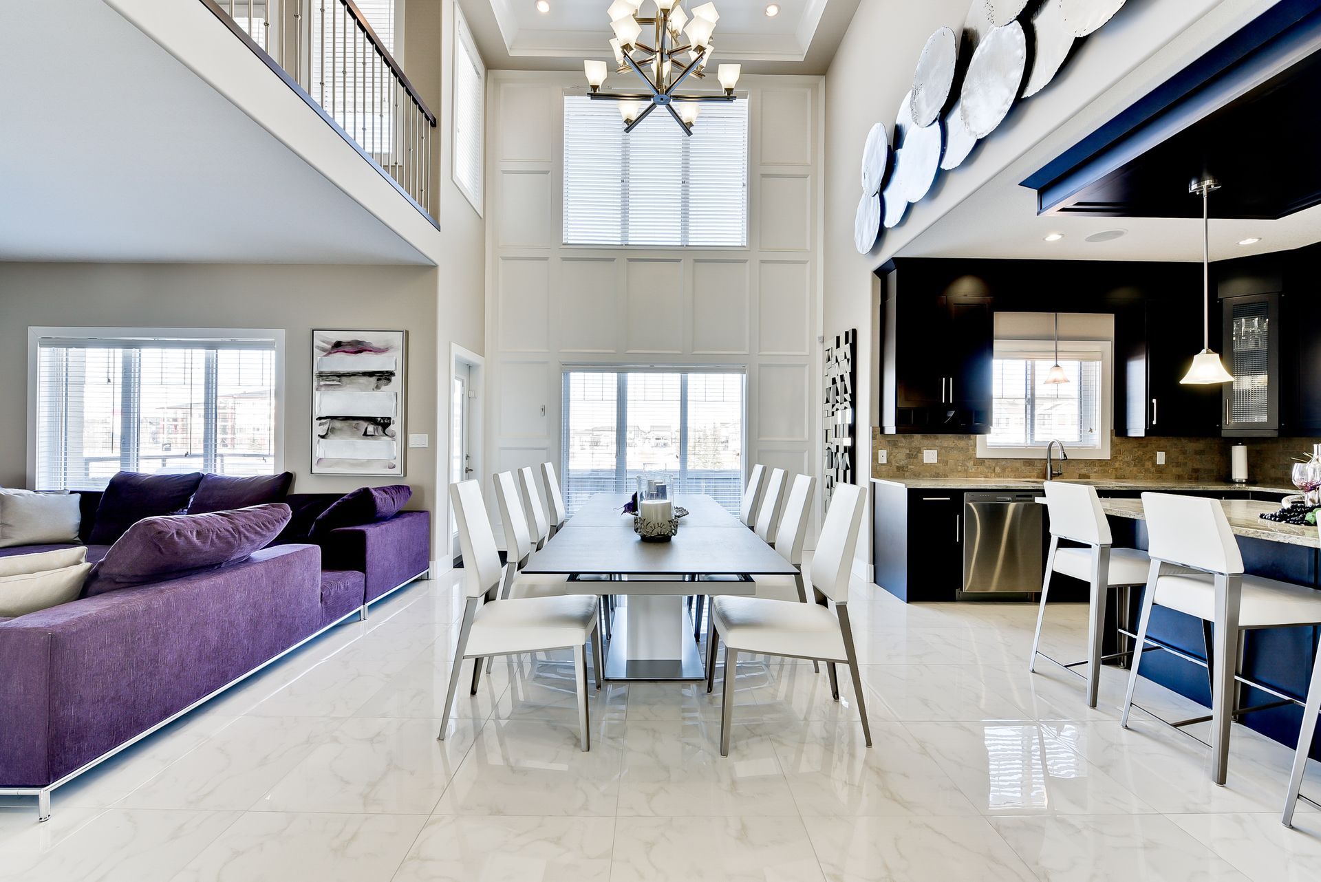 A modern, open-concept dining and living area with a purple sofa, long dining table, and dark kitchen. High ceilings and large windows.
