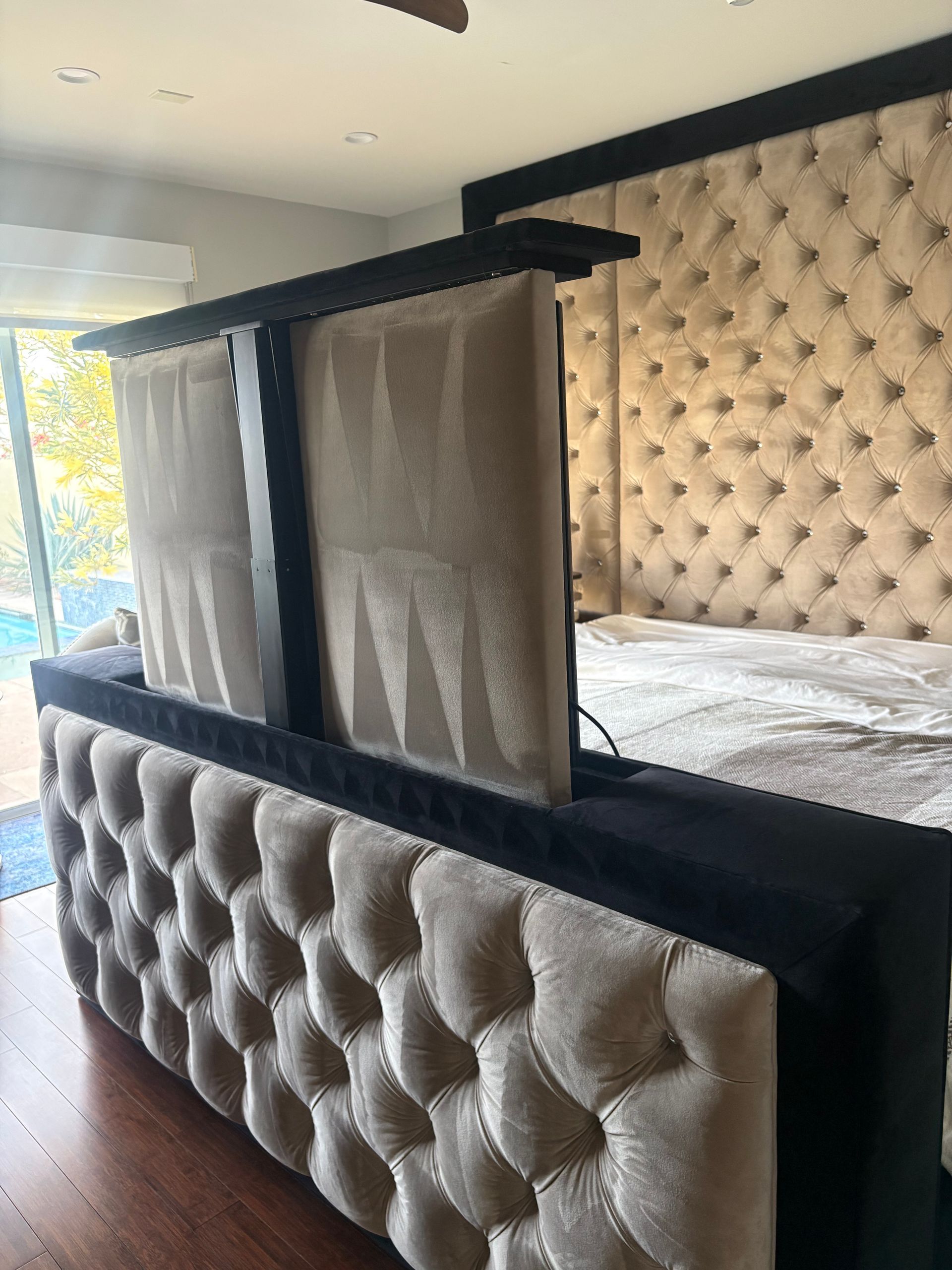 Beige button-tufted bed with a pop-up TV. Black frame. Sliding glass door in the background.