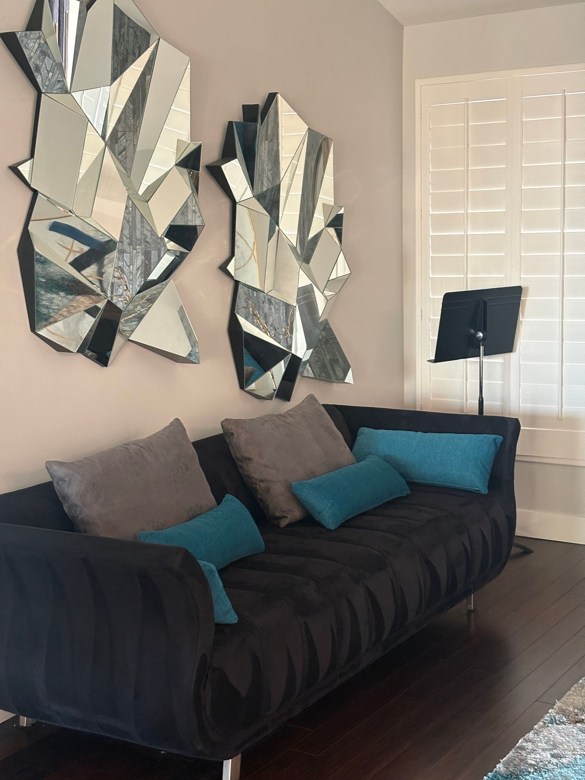 Black couch with teal and gray pillows, two geometric mirrors on wall, music stand by window.
