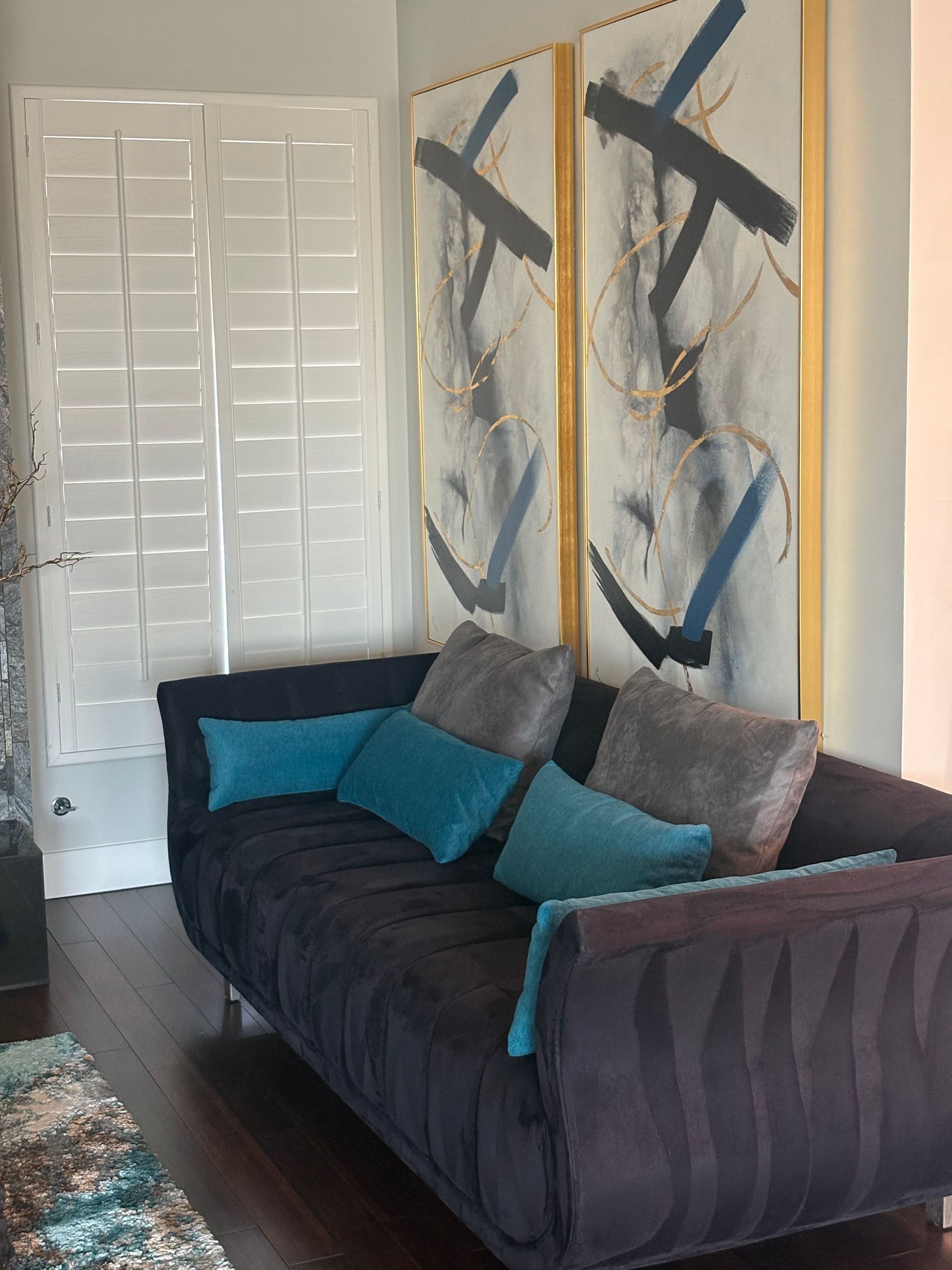 Living room with a dark sofa, teal and gray pillows, two abstract paintings, and white shutters.