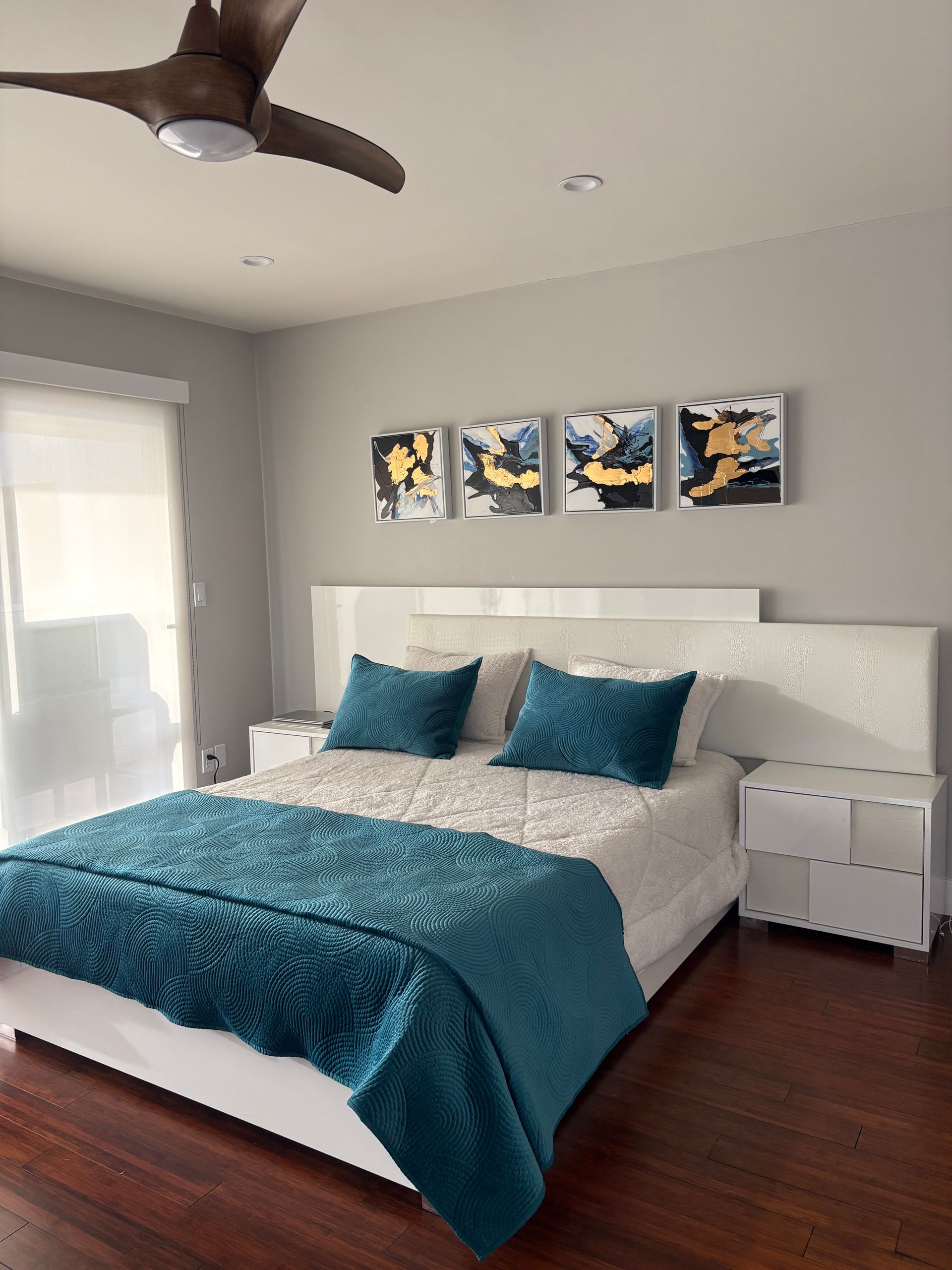 Bedroom with a white bed, teal blanket and pillows, dark wood floor, and abstract art.
