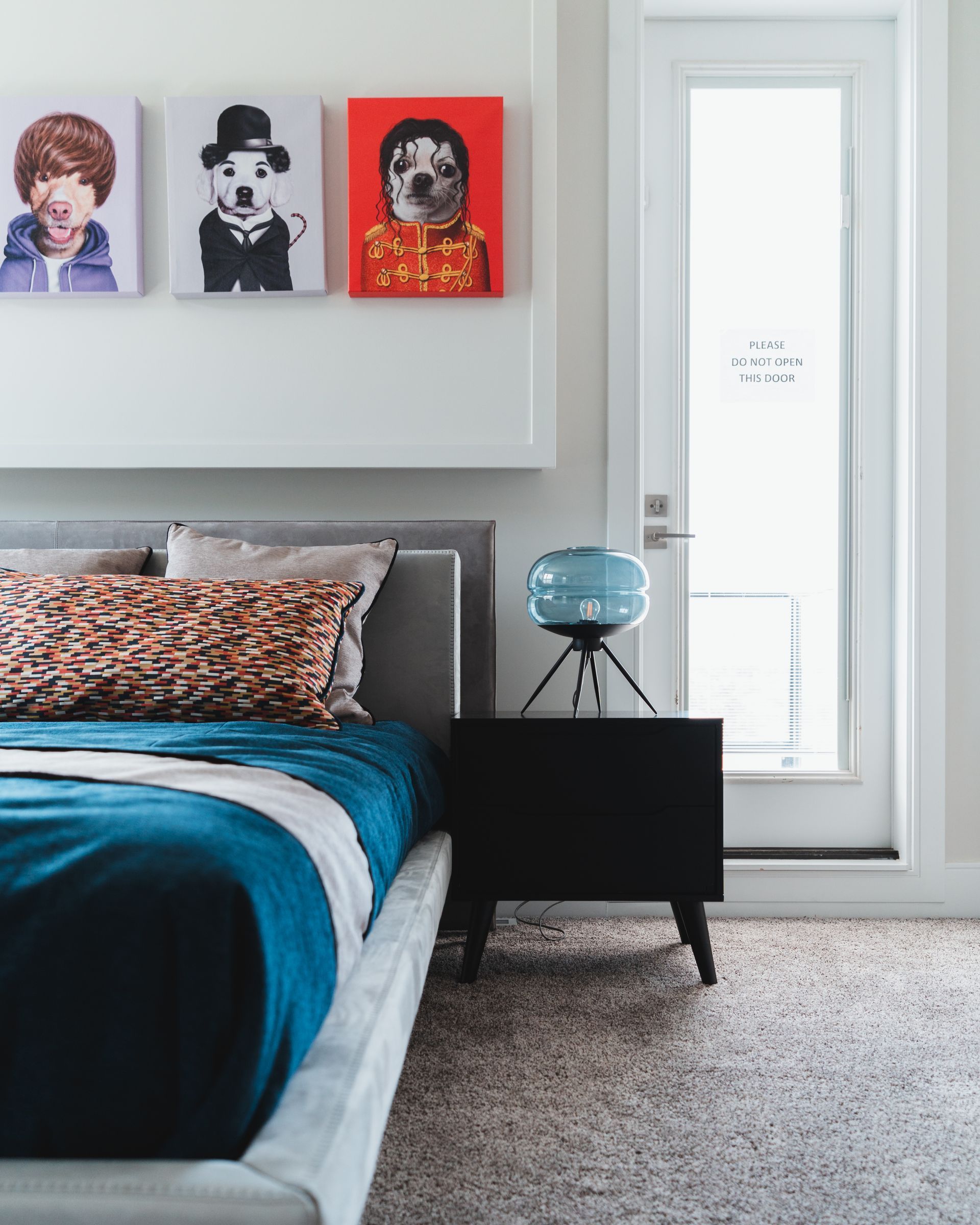 Bedroom with a bed, art, and nightstand. The bed has a blue blanket and patterned pillows.  Artwork above the bed depicts stylized portraits. A blue lamp sits on the nightstand.