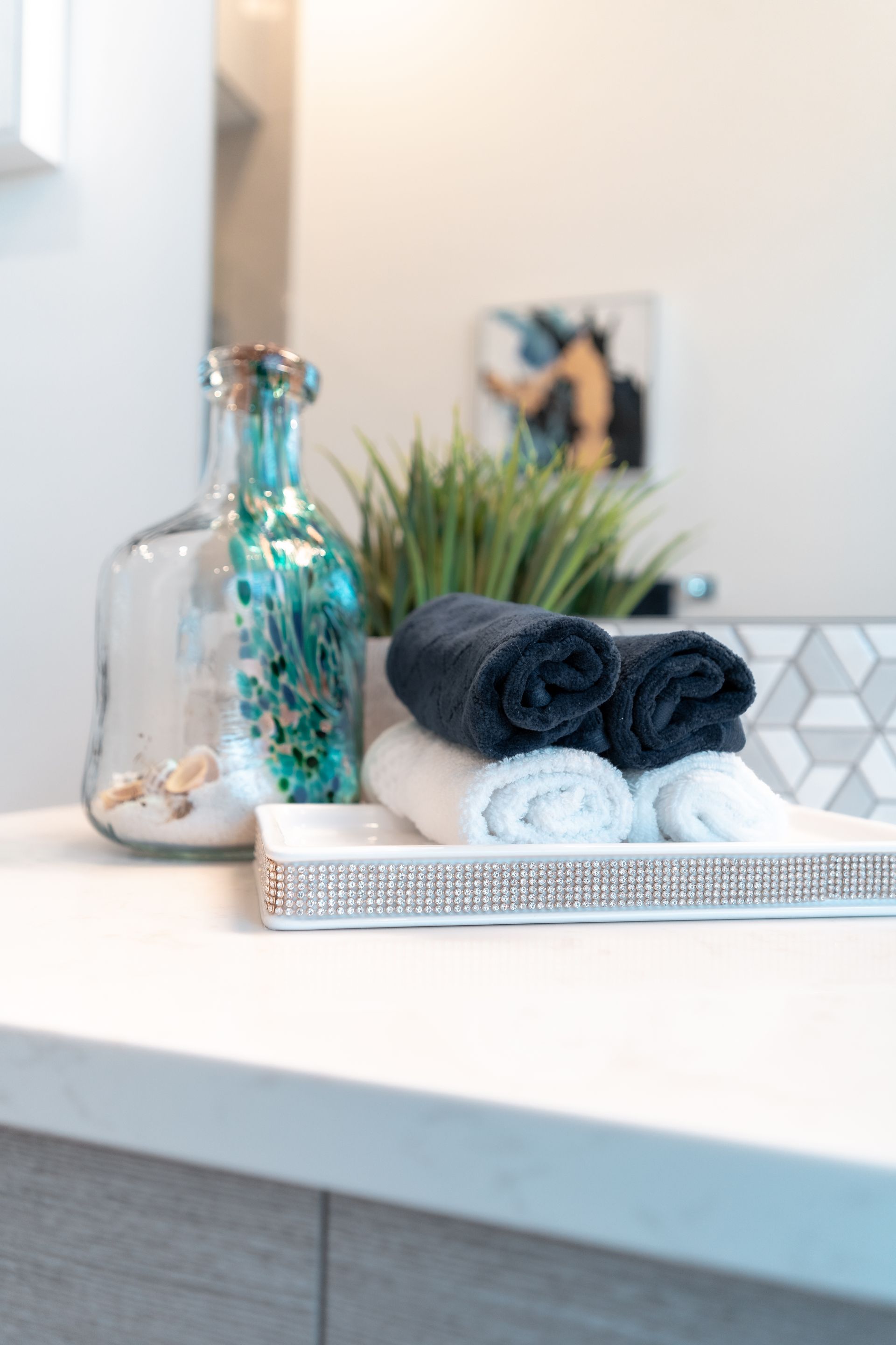 A bathroom counter with a tray of towels and a bottle of seashells.