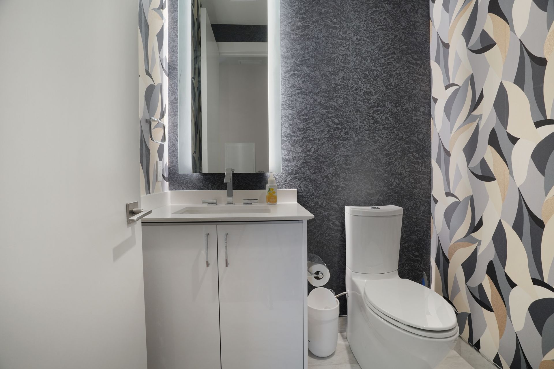 Small modern bathroom with white vanity, toilet, and patterned shower curtain; walls are white and speckled black.