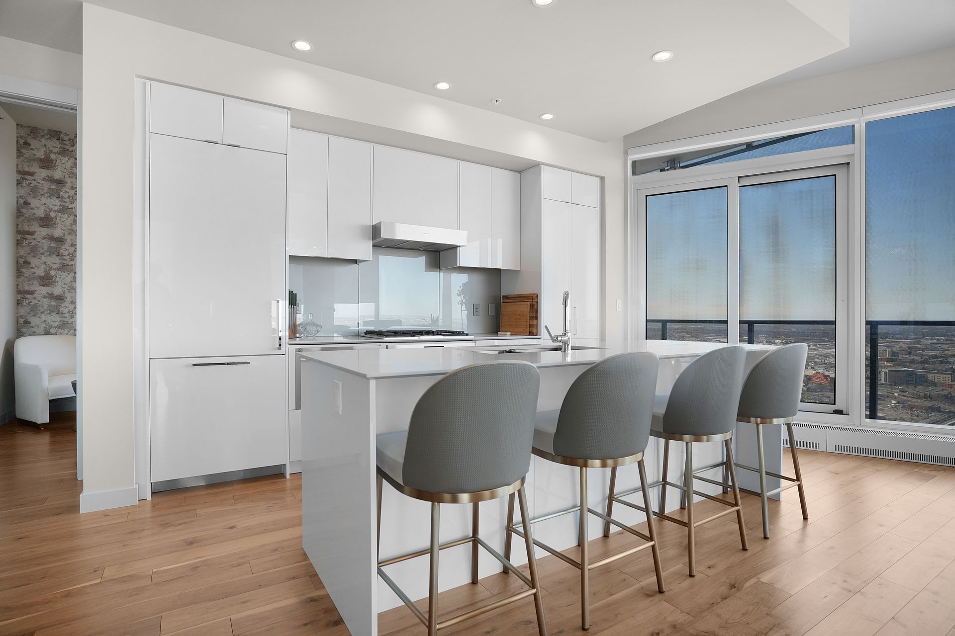 Modern white kitchen with an island and grey bar stools, overlooking a cityscape through large windows.