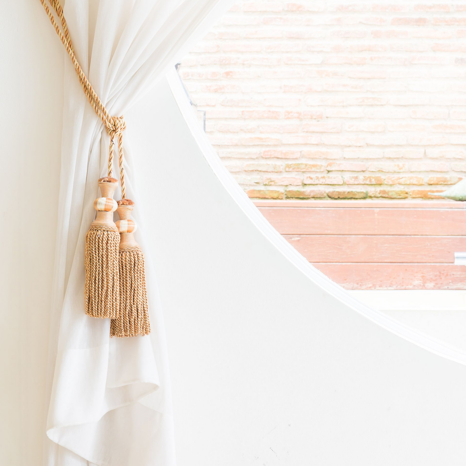 White curtain draped open with gold tassel tieback, revealing a window.