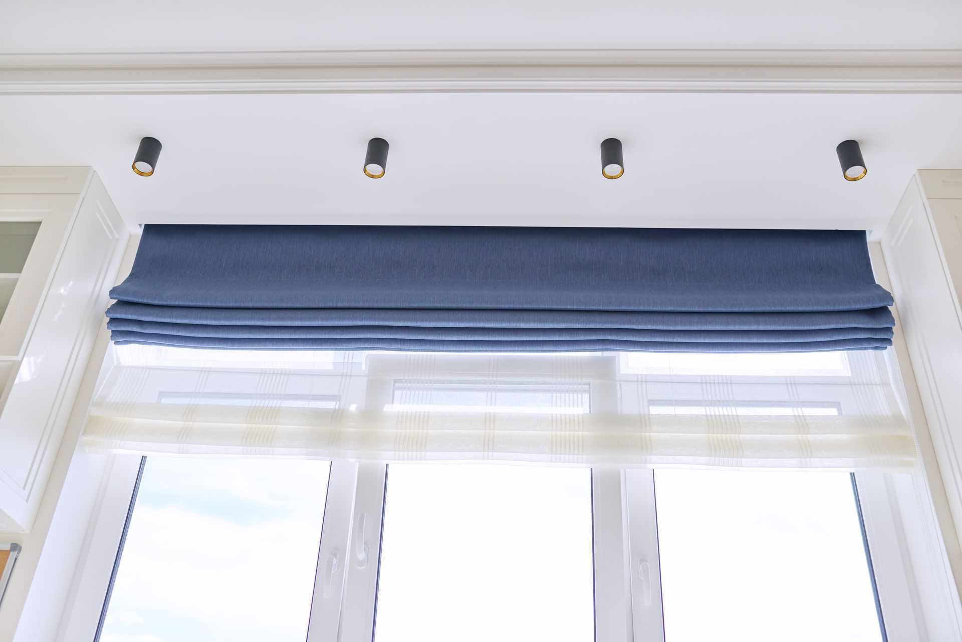 Blue roman shade over a sheer white curtained window. Recessed lights are above.