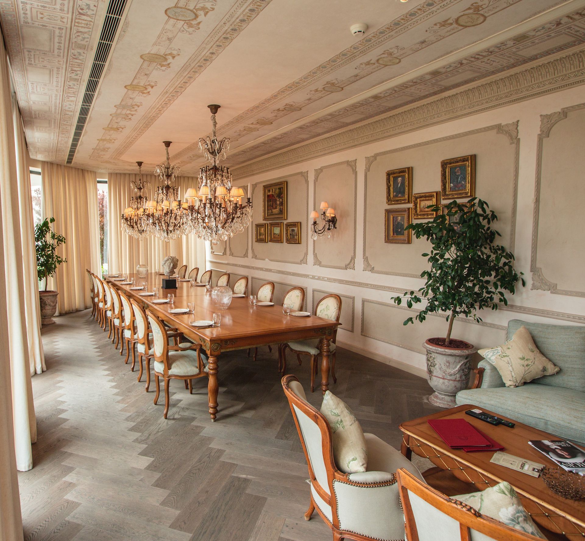 Elegant dining room with a long wooden table, chandeliers, ornate wall panels, and large windows