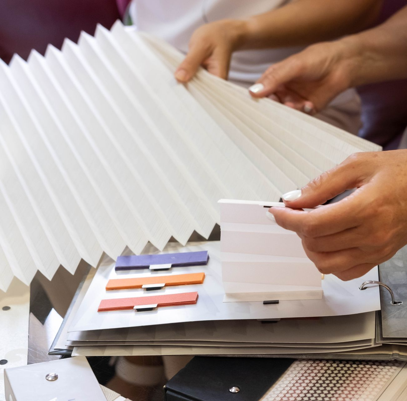 Hands examining pleated blinds samples with color swatches on a table.