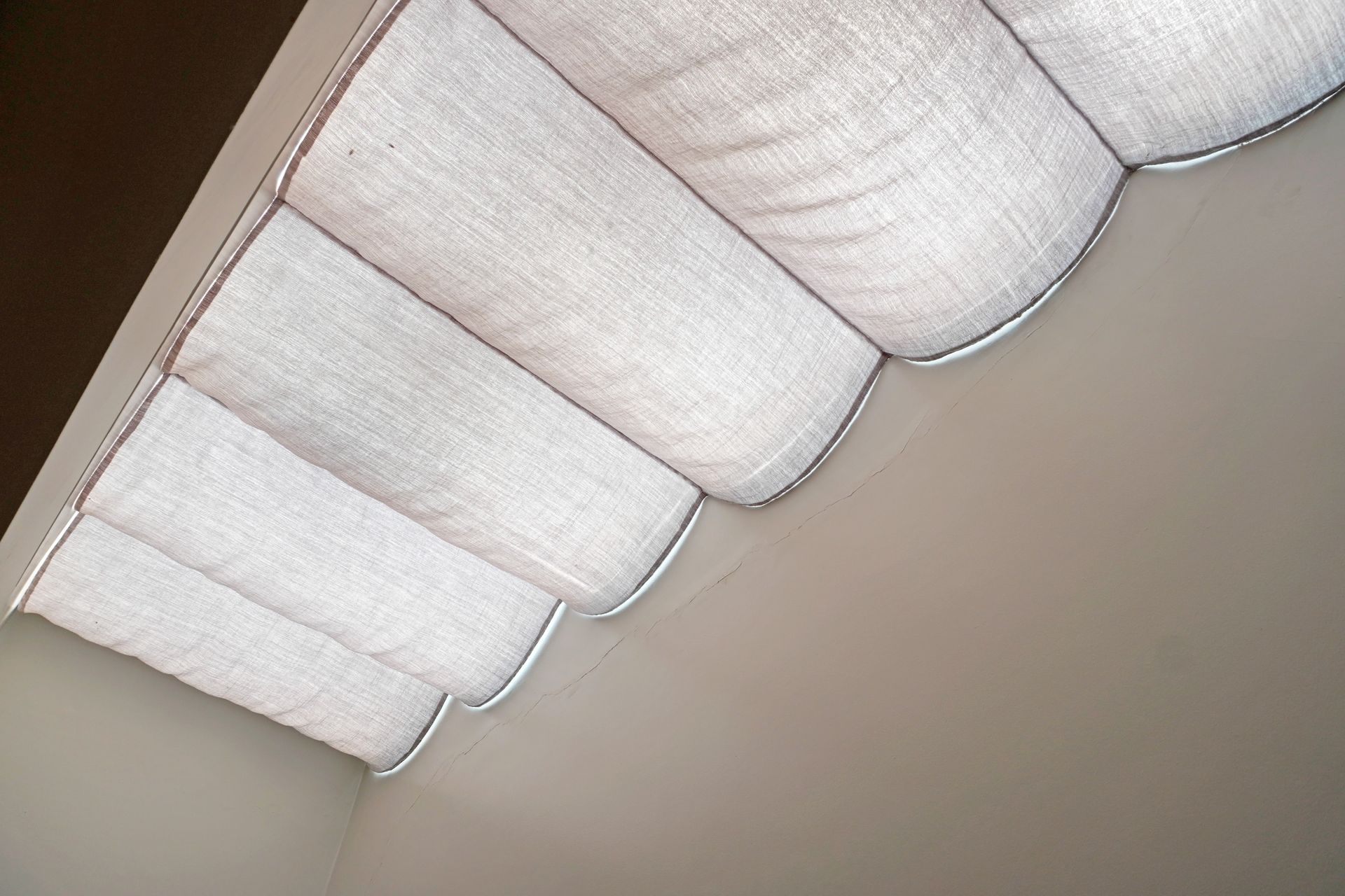 White fabric window shade, with scalloped bottom edge, angled against a neutral wall.