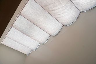 White fabric window shade, with scalloped bottom edge, angled against a neutral wall.