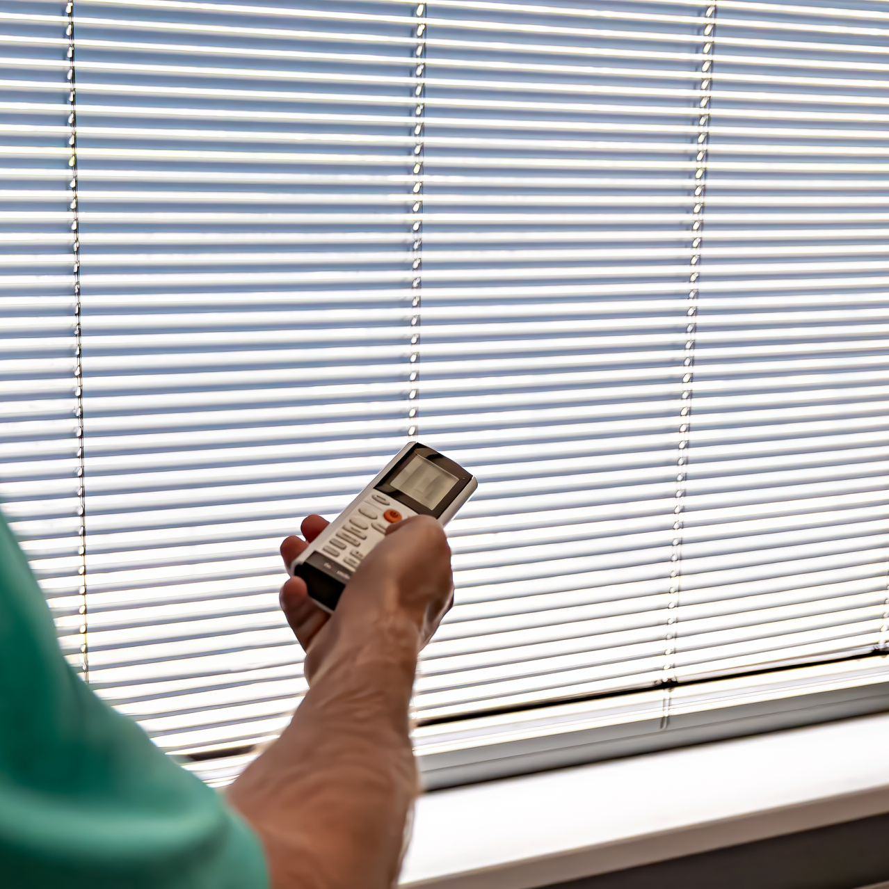 Person holding a remote control adjusting horizontal blinds in a window; bright daylight.