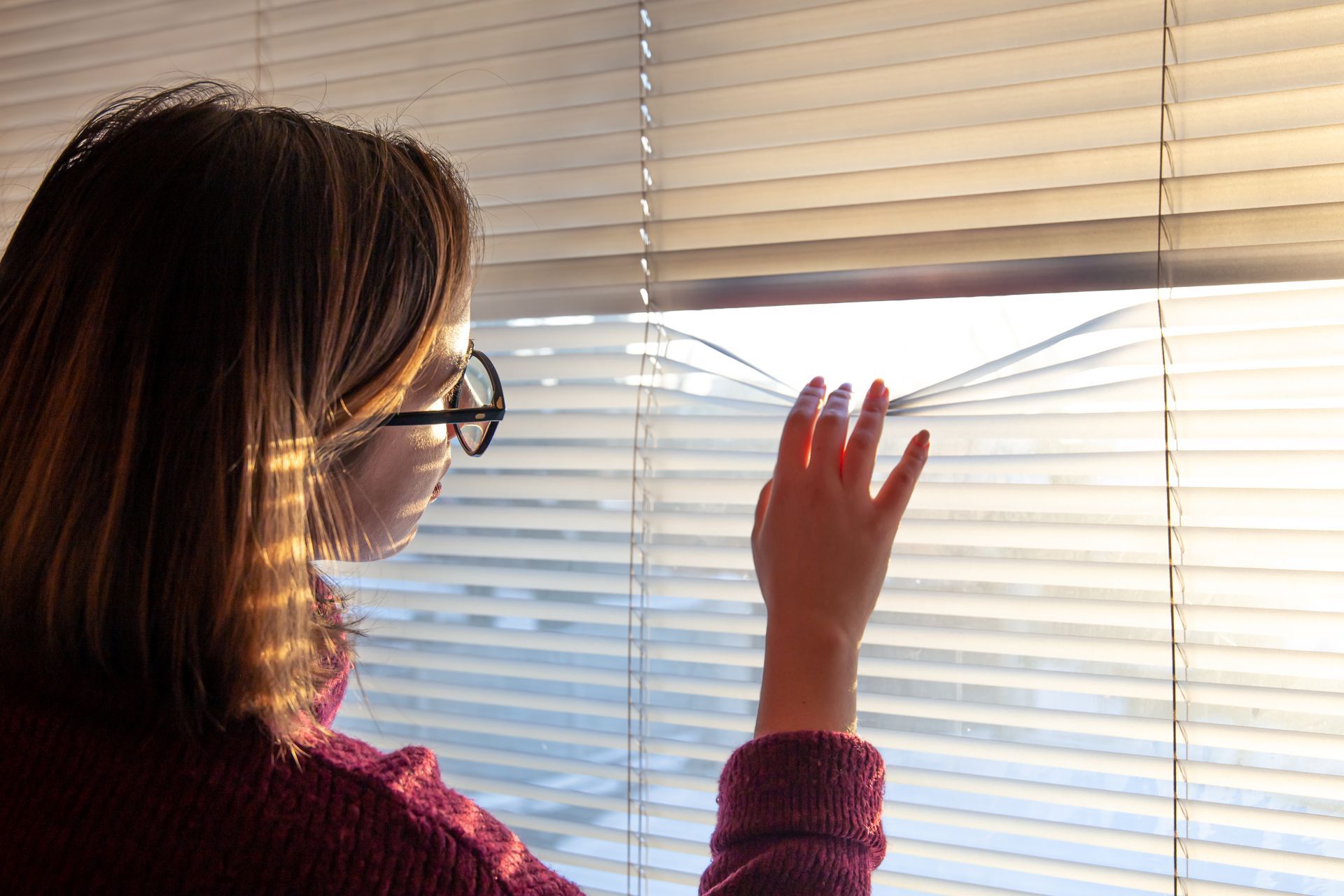 Woman with glasses peeking through window blinds, sunlight streaming in.