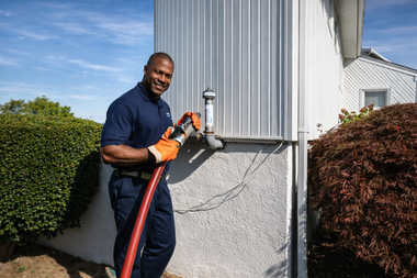 A professional in a navy uniform attaches a red fuel hose to an outdoor home heating oil fill pipe.