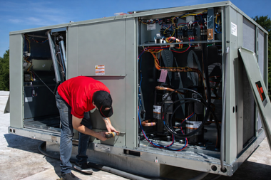 Technician in a red shirt repairing a large rooftop HVAC unit on a sunny day.
