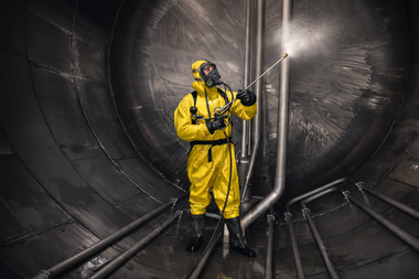 A person in a yellow protective hazmat suit uses a high-pressure spray wand to clean the metal interior of a large tank.