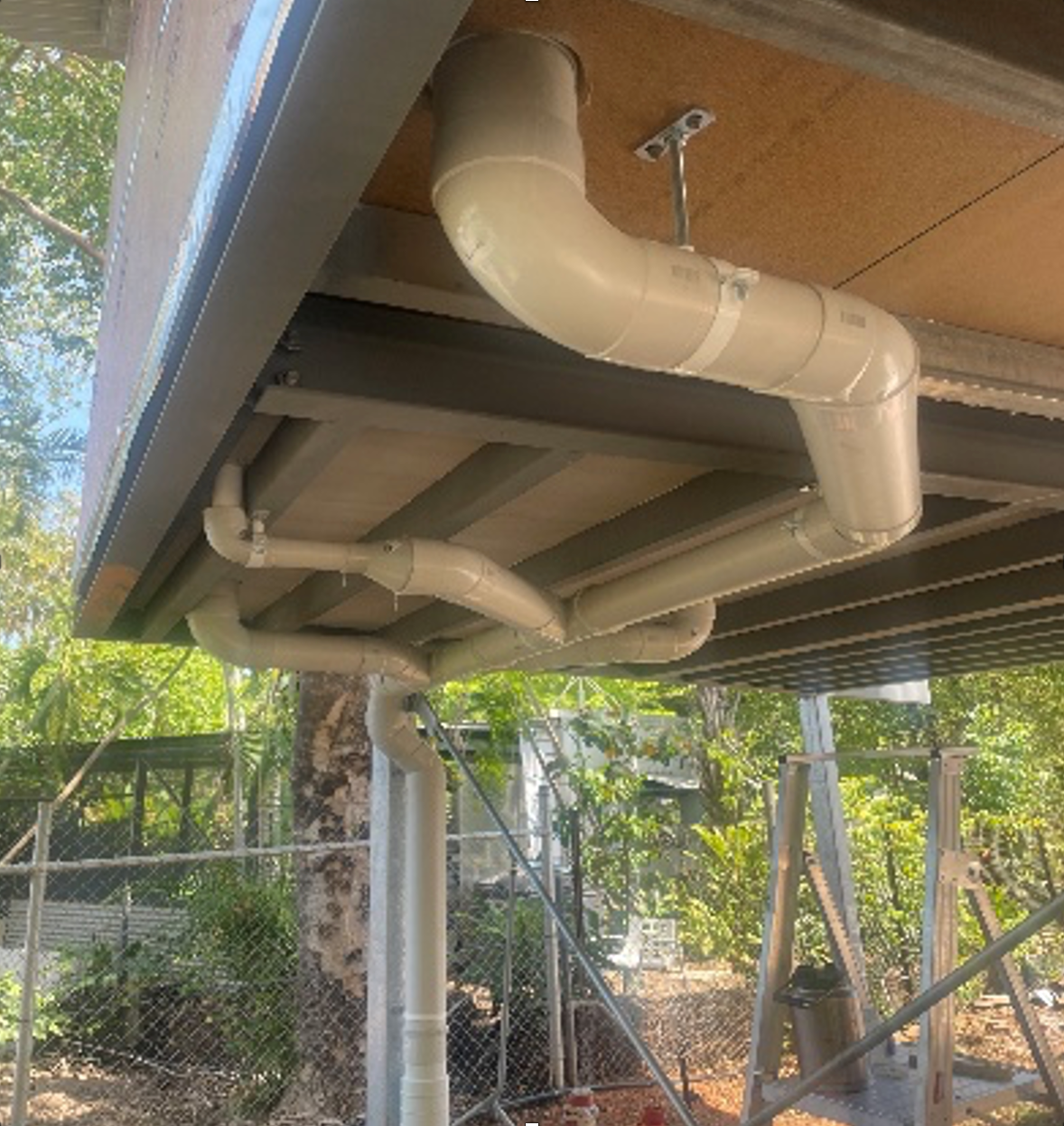 PVC pipes under a building, leading outside; daylight setting, trees in background. — Ausplumb NT in Winnellie, NT