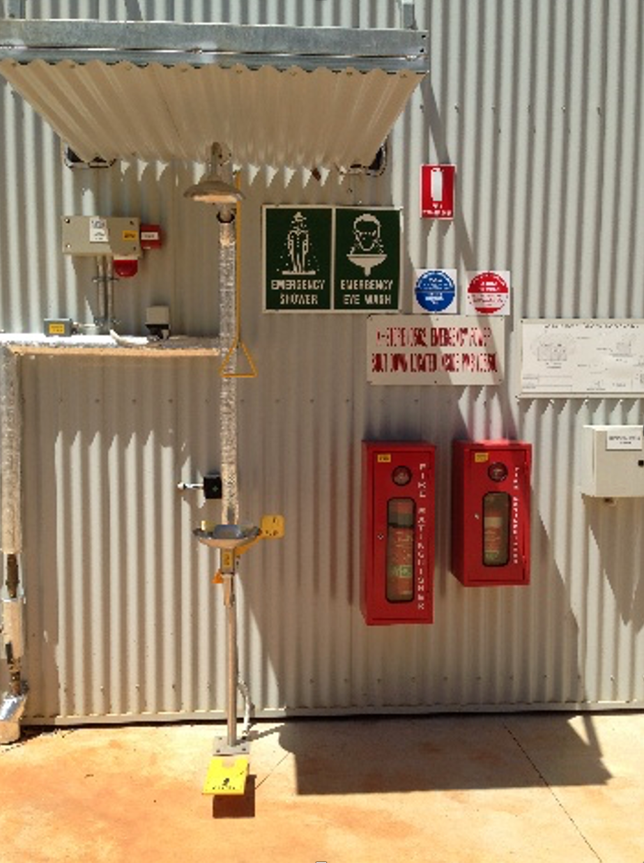 Emergency shower, eyewash station, fire hoses, and safety signs on a corrugated metal building. — Ausplumb NT in Winnellie, NT