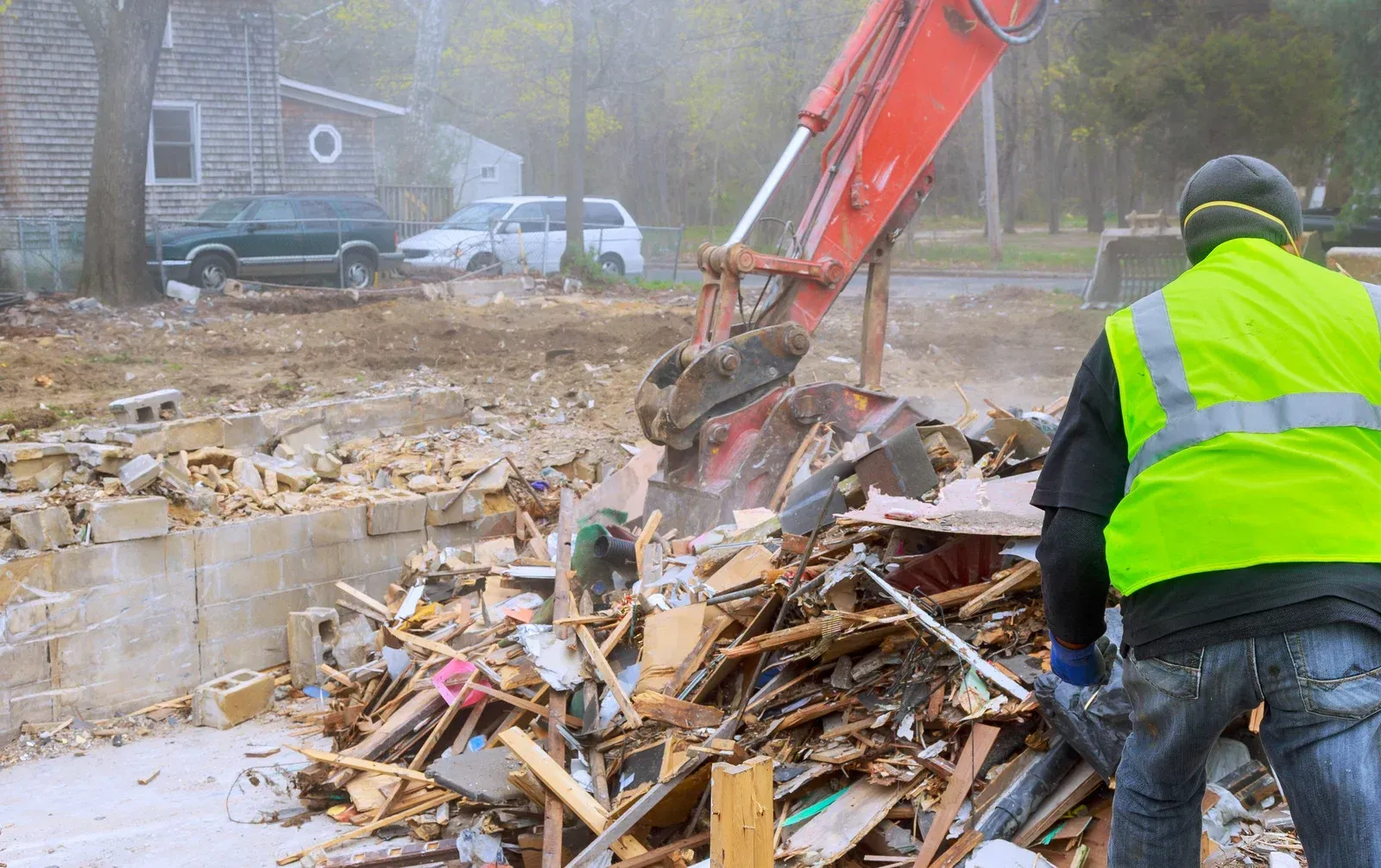 Demolition site: Excavator with claw arm, worker in safety vest, debris pile, residential setting.