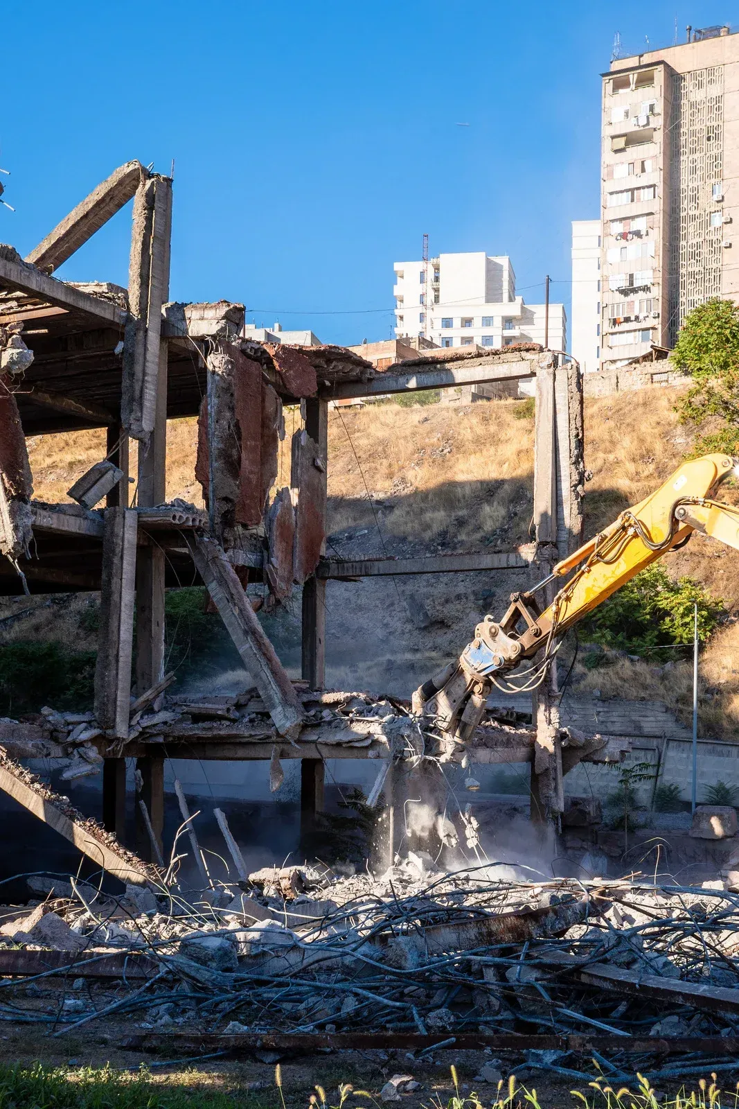 Demolition of a building with a yellow excavator against a hillside. Debris and dust fill the air on a sunny day.