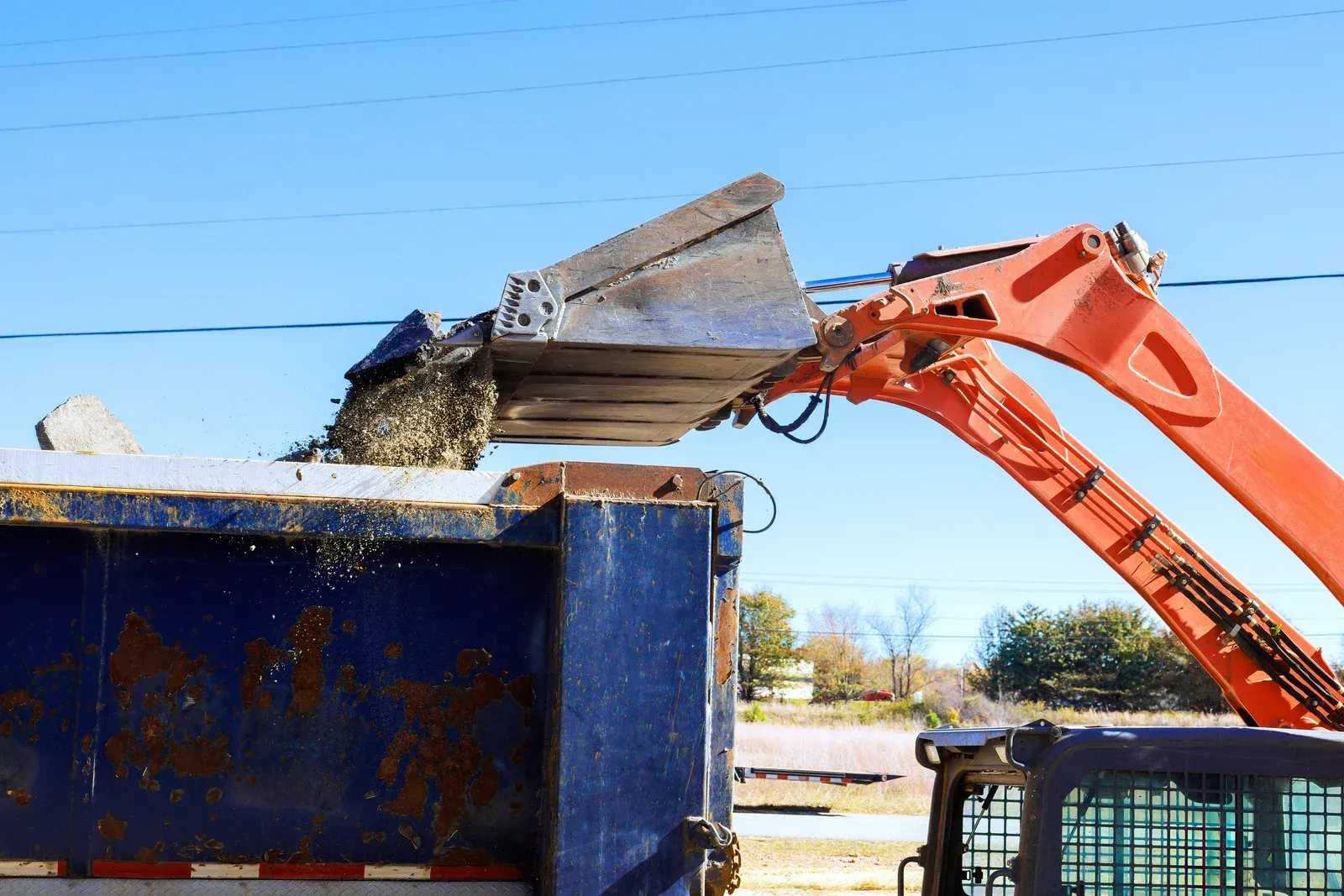 Orange excavator dumping debris into a blue dump truck against a clear sky.