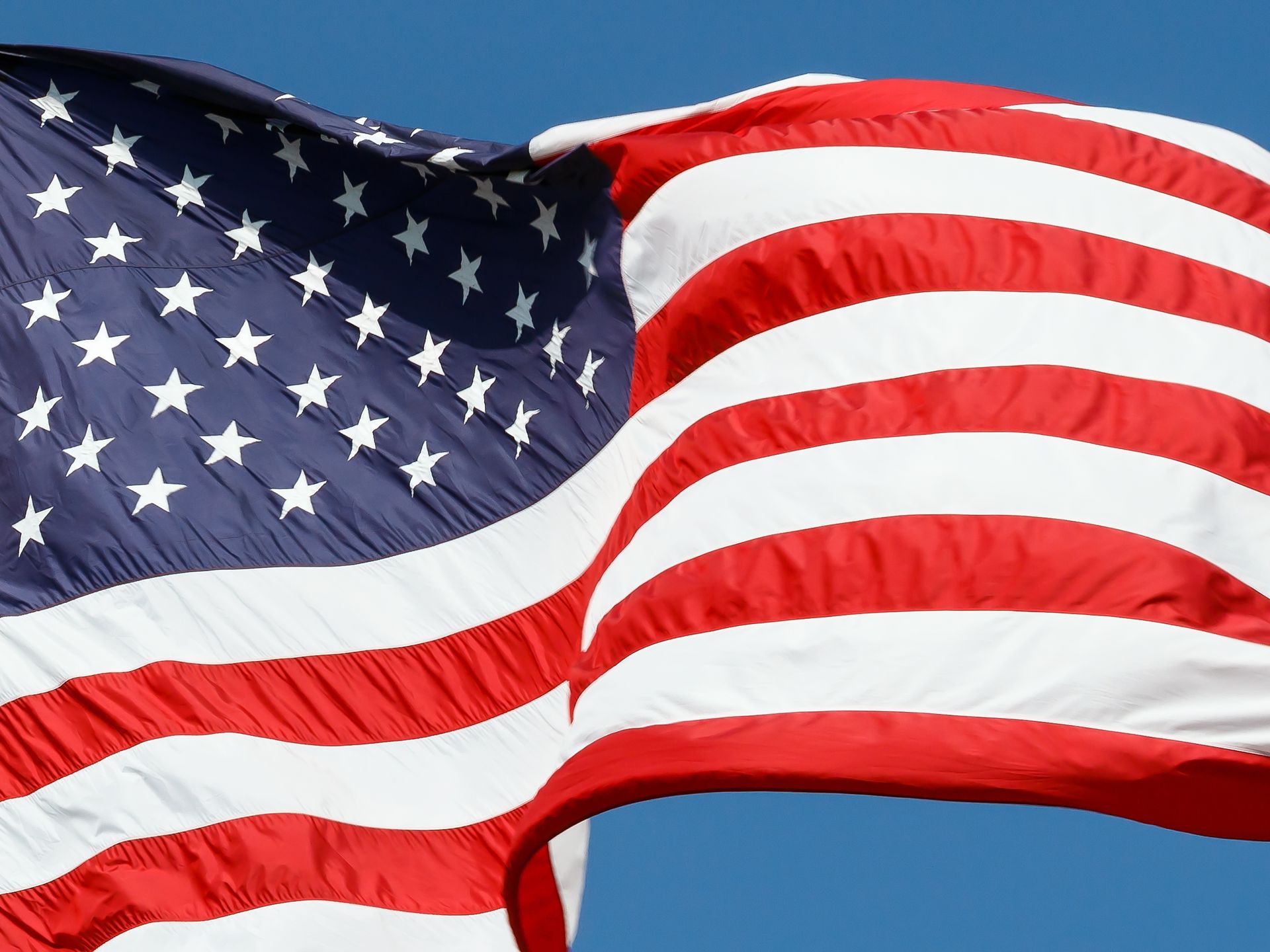 American flag waving in front of a blue sky, red and white stripes, stars in the blue field.