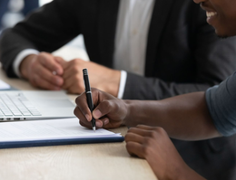 Hands signing a document; a small house model sits nearby on a wooden table. Hands signing a document; a small house model sits nearby on a wooden table.