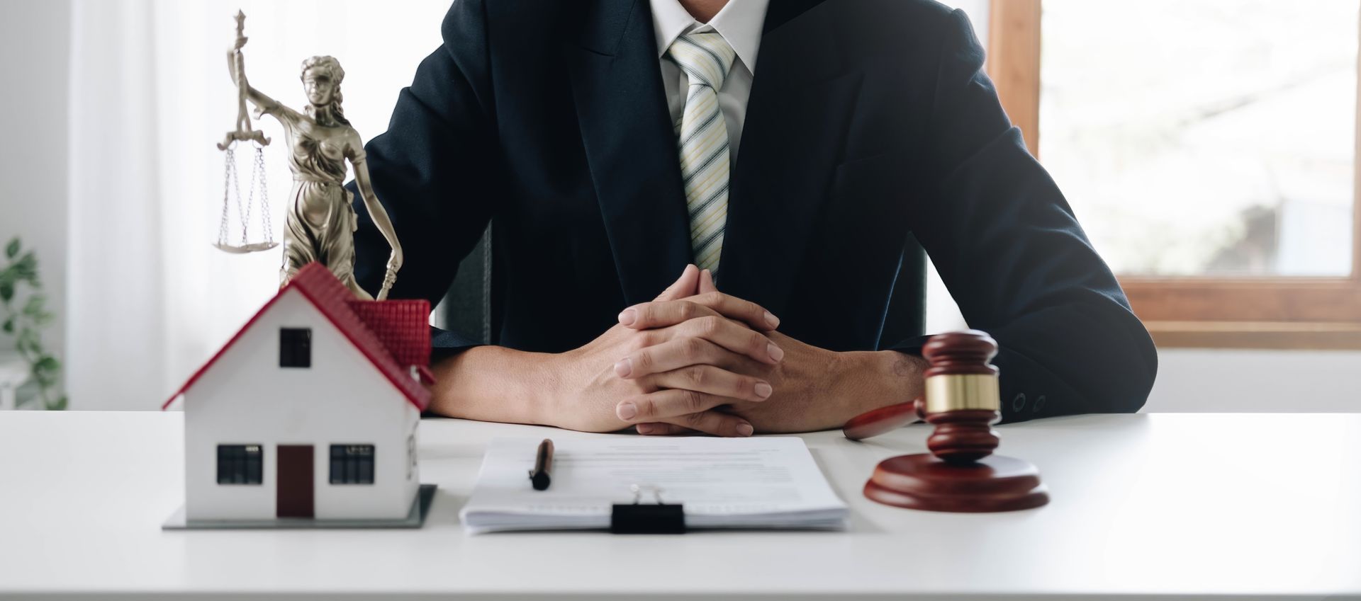 A lawyer in a suit sits at a desk with a gavel, statue of Lady Justice, a toy house, and paperwork. A lawyer in a suit sits at a desk with a gavel, statue of Lady Justice, a toy house, and paperwork.