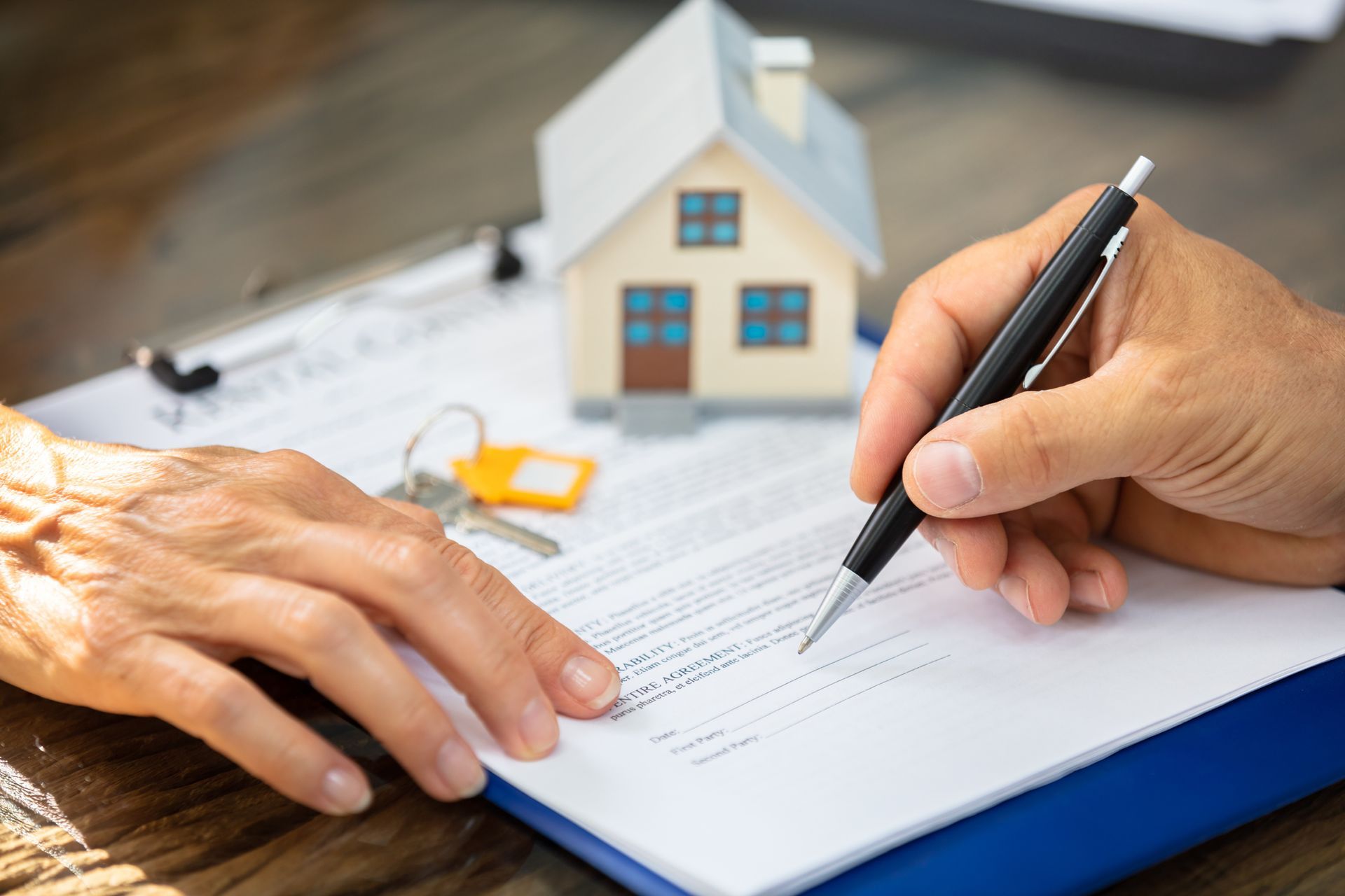 Person signing a document, with a toy house and keys on a table.