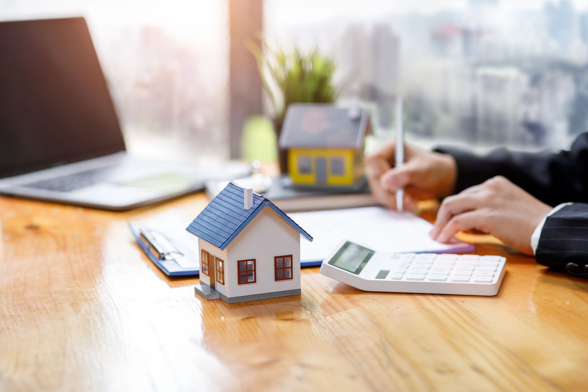 A person writing on a document with a miniature house, laptop, and calculator on a wooden desk.