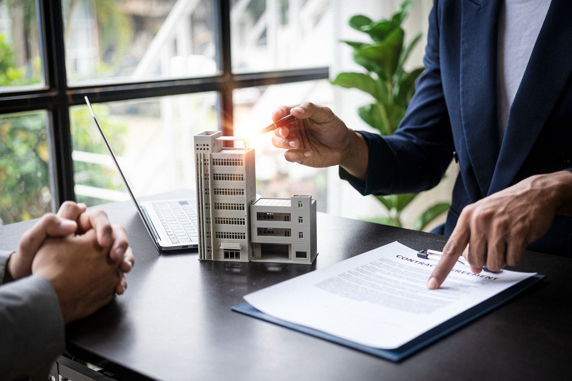 Person pointing at contract, small building models, laptop on table, bright window background.