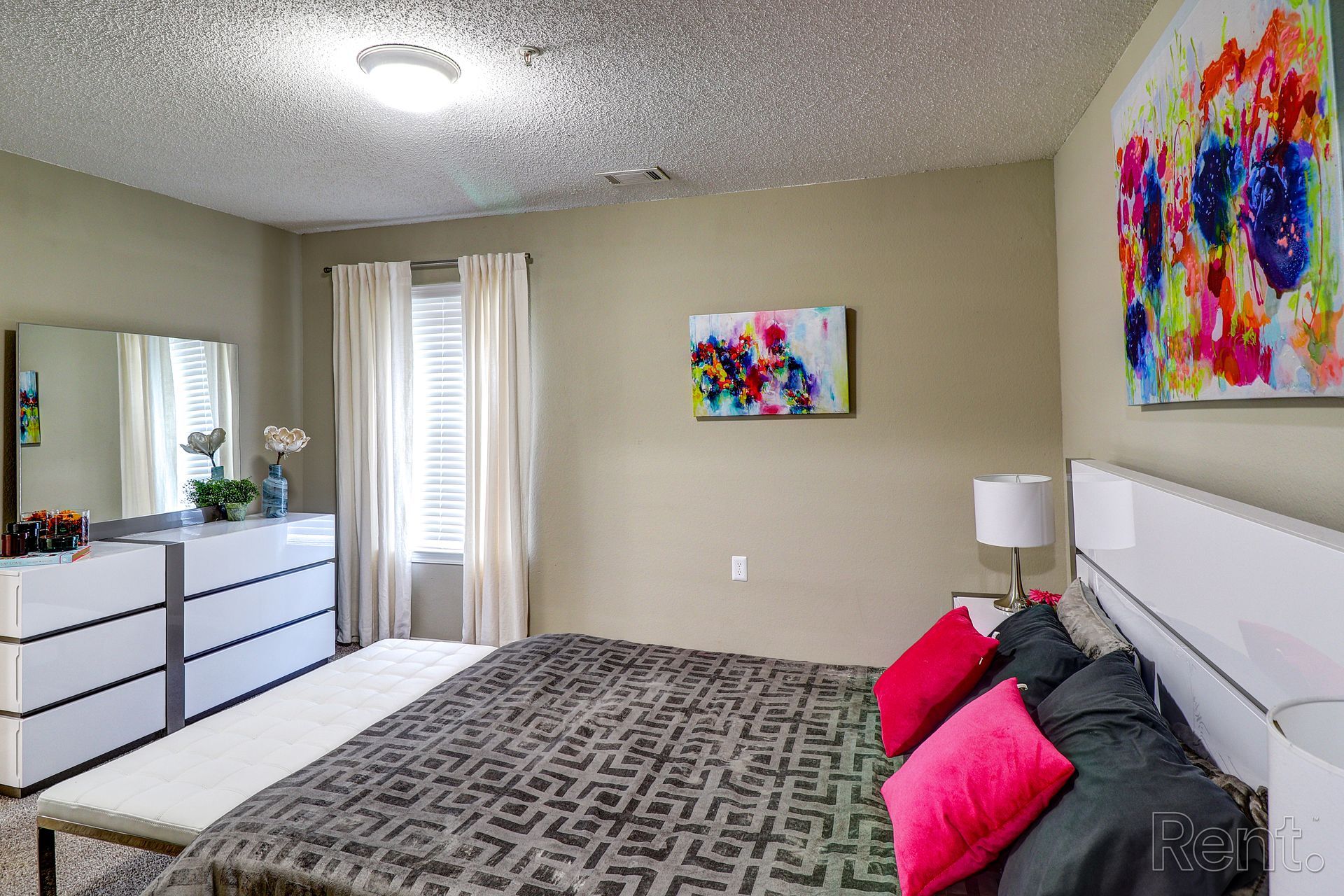 A bedroom with a large bed , dresser , mirror and painting on the wall.