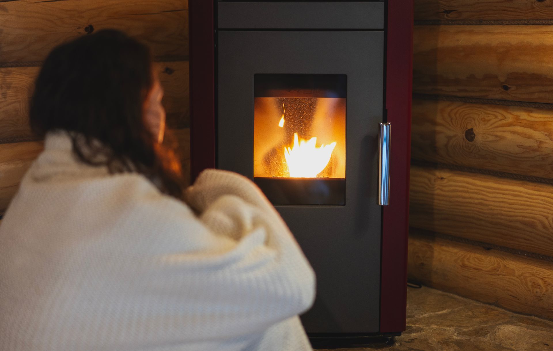 Woman in white robe sits near lit stove in wooden cabin.