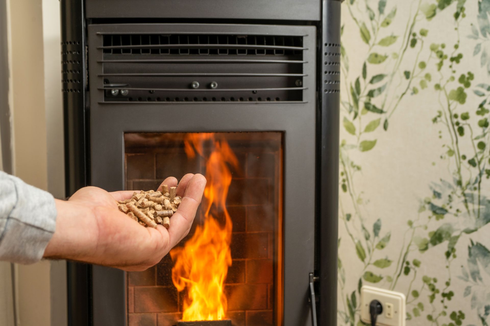 Handful of wood pellets being held over a burning pellet stove, near floral wallpaper and a wall outlet.
