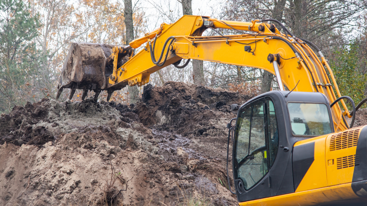 Yellow excavator on muddy terrain, digging earth under a cloudy sky.