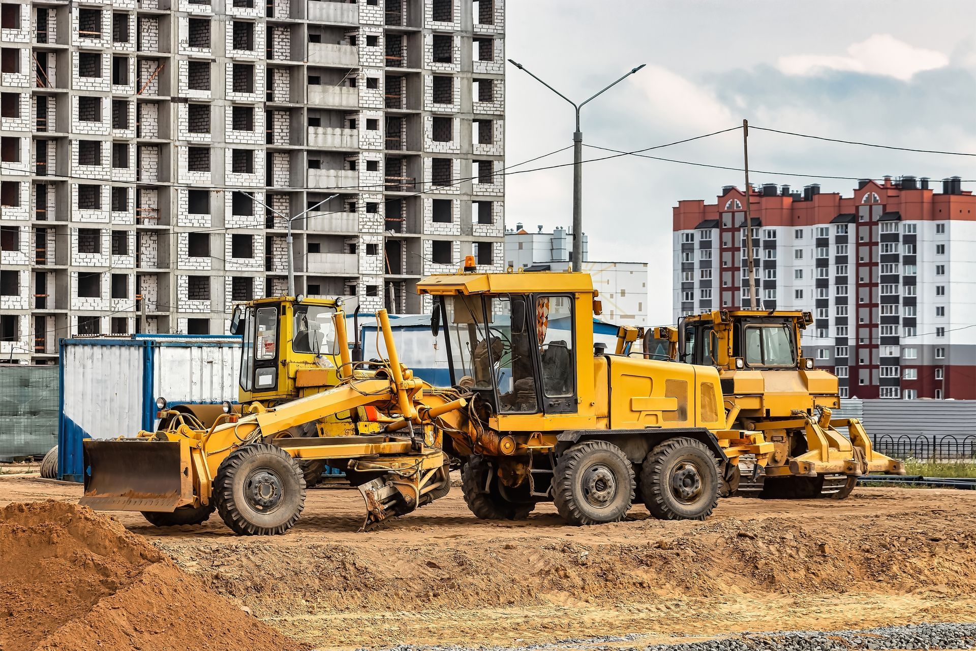 Yellow construction graders on a dirt lot in front of a partially built high-rise building.