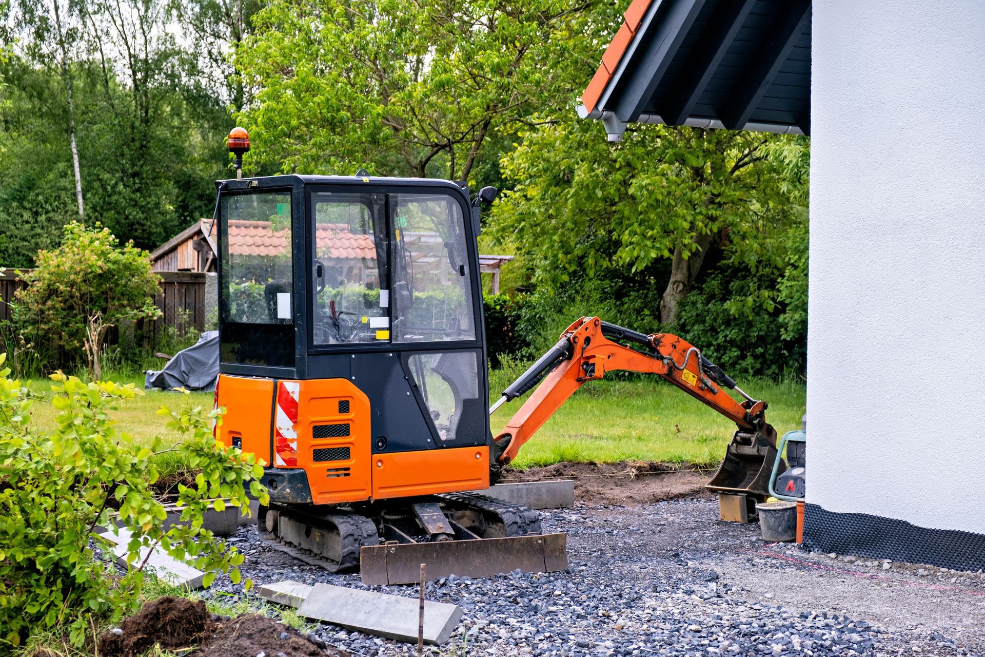 Orange mini excavator parked next to a house with an attached black-roofed overhang, gravel area.