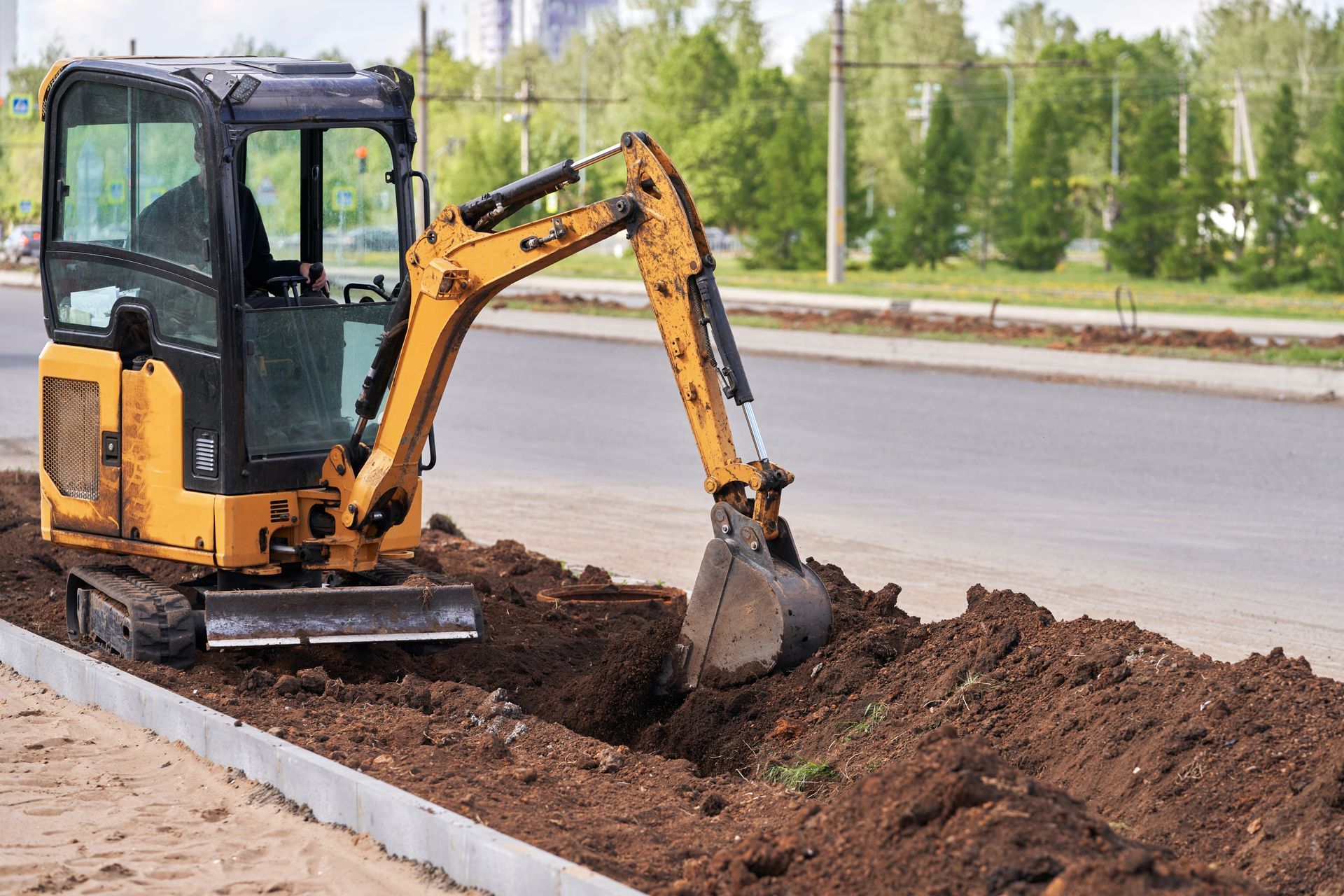 Yellow mini-excavator digging a trench alongside a road.
