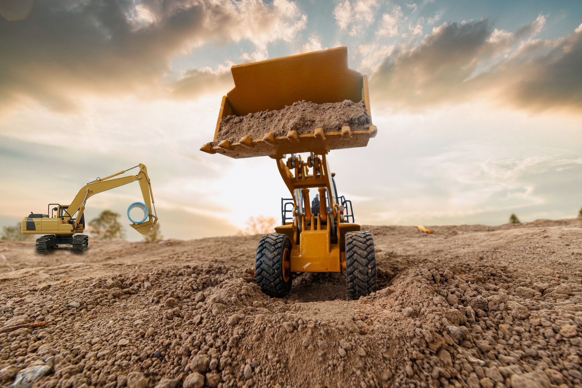 Yellow front loader and excavator working on dirt ground under a cloudy sky.